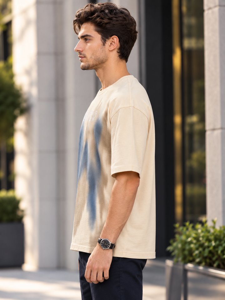 A man wearing a cream-colored short-sleeve t-shirt with a blue abstract graphic print, standing in an outdoor urban setting by mentor.