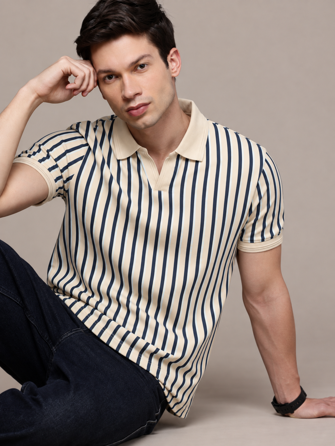 A man wearing a cream and navy blue vertically striped short-sleeve polo shirt with dark jeans, posing against a solid beige studio background.