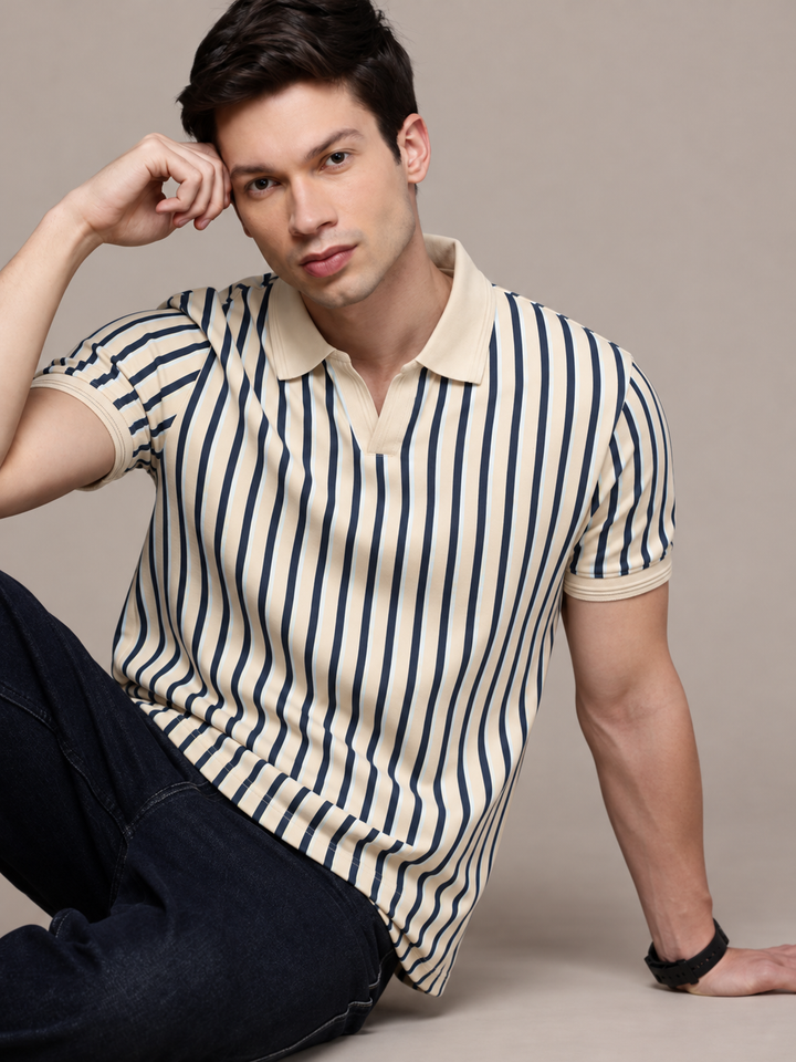 A man wearing a cream and navy blue vertically striped short-sleeve polo shirt with dark jeans, posing against a solid beige studio background.