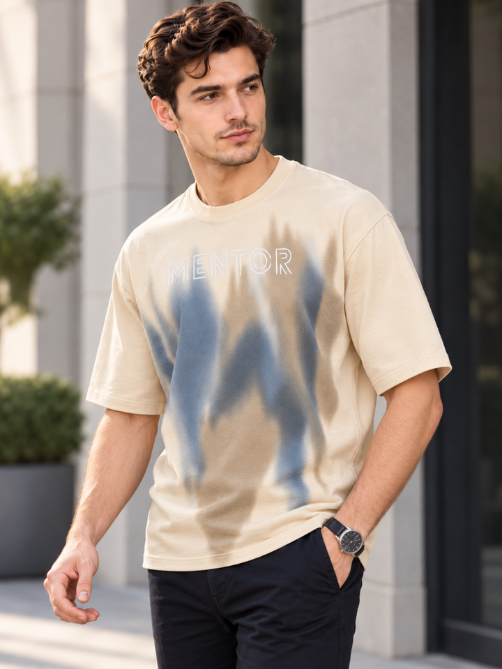 A man wearing a beige short-sleeve t-shirt with a blue abstract graphic and the word 'MENTOR' printed on the chest, paired with dark pants, standing outdoors in front of a modern building.