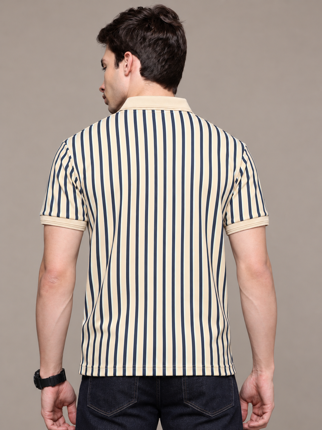 A man viewed from behind wearing a short-sleeve polo shirt with vertical navy blue and cream stripes, paired with dark jeans, against a solid taupe background.