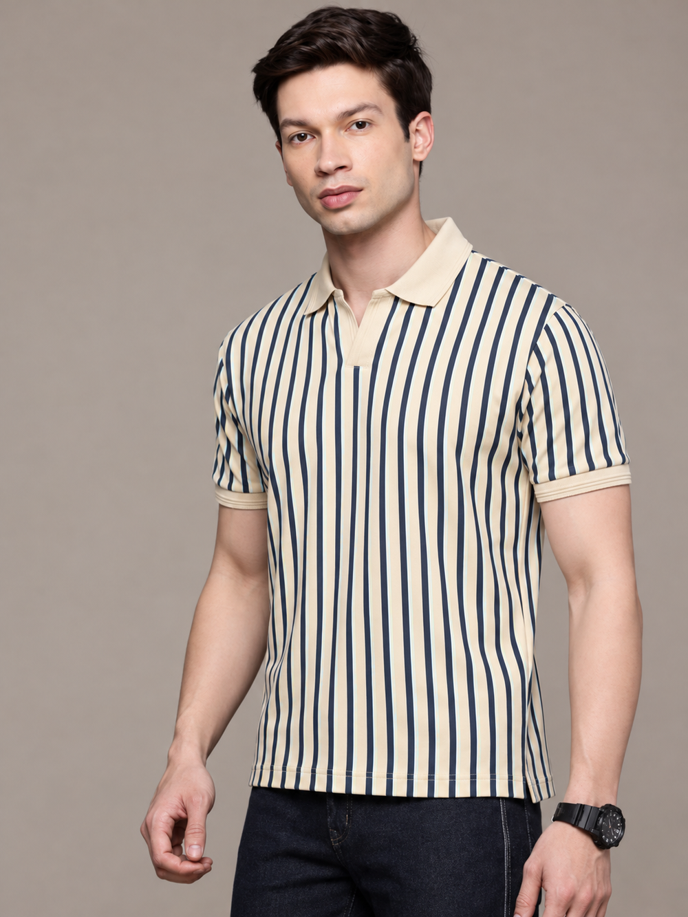 A man wearing a short-sleeve polo shirt with vertical navy blue and cream stripes, paired with dark jeans, standing against a solid taupe background.