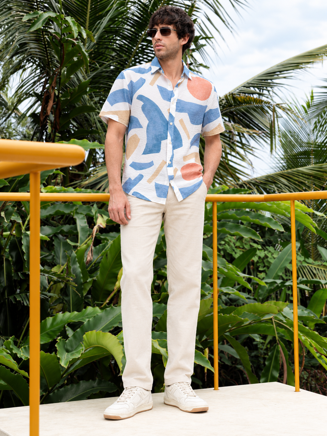 A man wearing a short-sleeve button-up shirt with a blue, beige, and orange abstract patchwork pattern, paired with cream trousers and white sneakers, standing outdoors in front of a yellow railing and lush tropical greenery.