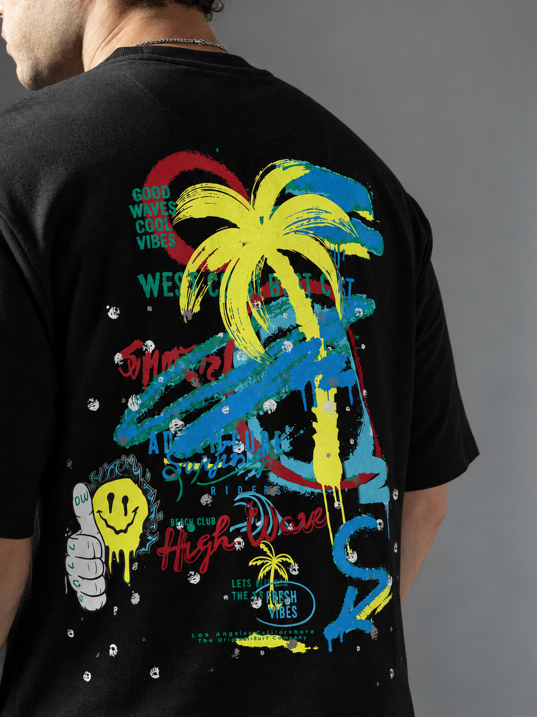 A man wearing a black t-shirt, viewed from the back. The shirt features a large, vibrant graphic print with a yellow palm tree, blue and red paint strokes, graffiti-style text, and a yellow smiley face.