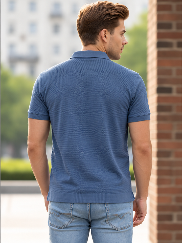 A man wearing a blue short-sleeve polo shirt and light blue jeans, shown from the back in an outdoor setting with a brick wall on the right.