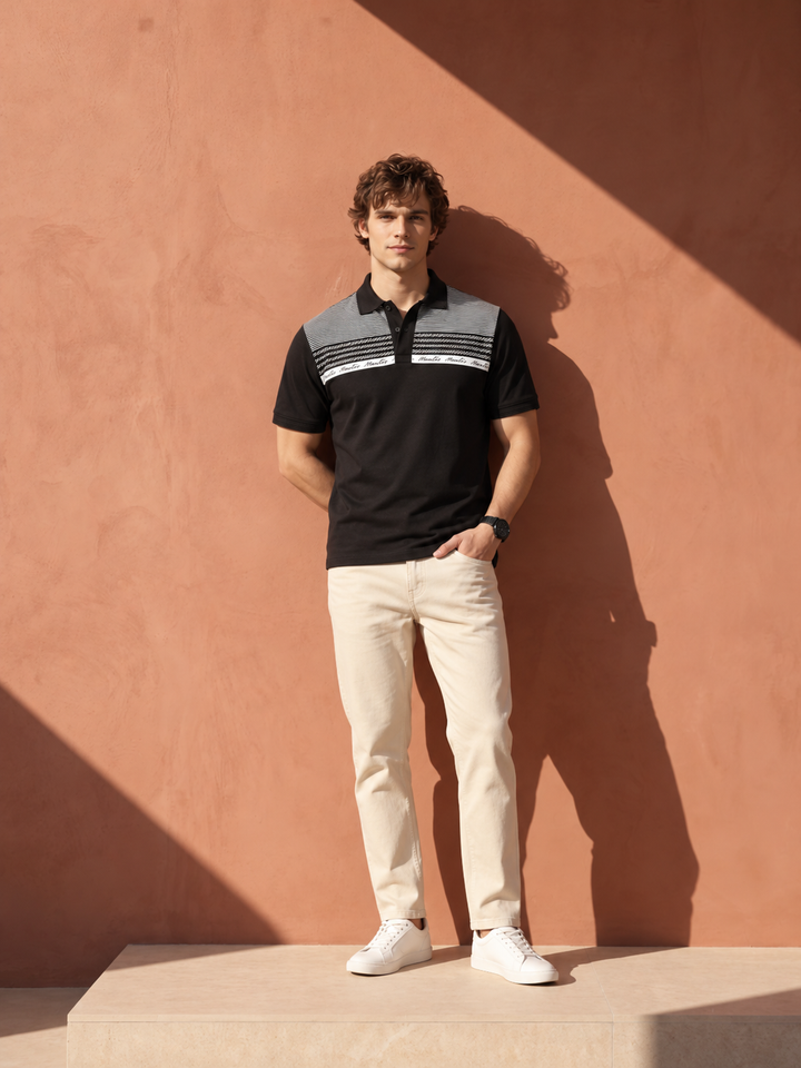 A man stands against a terracotta-colored wall wearing a black short-sleeve polo shirt with gray and white horizontal stripe details on the chest, paired with beige trousers and white sneakers.