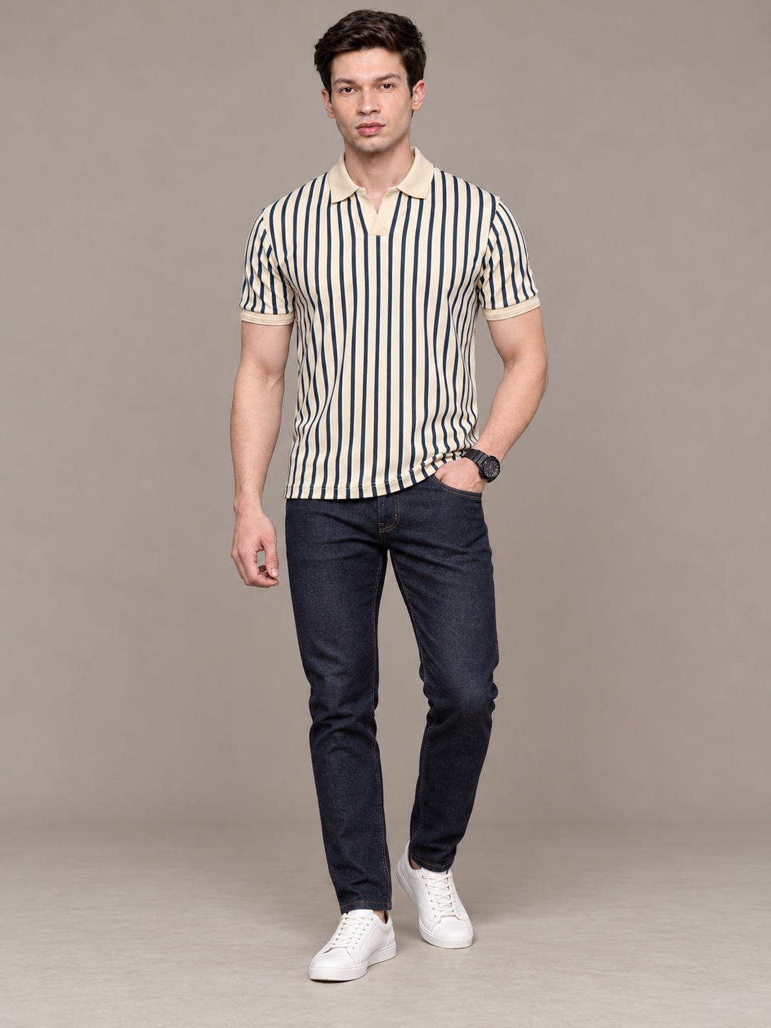 A man modeling a short-sleeve polo shirt with vertical navy and cream stripes, paired with dark blue jeans and white sneakers, against a solid beige studio background.
