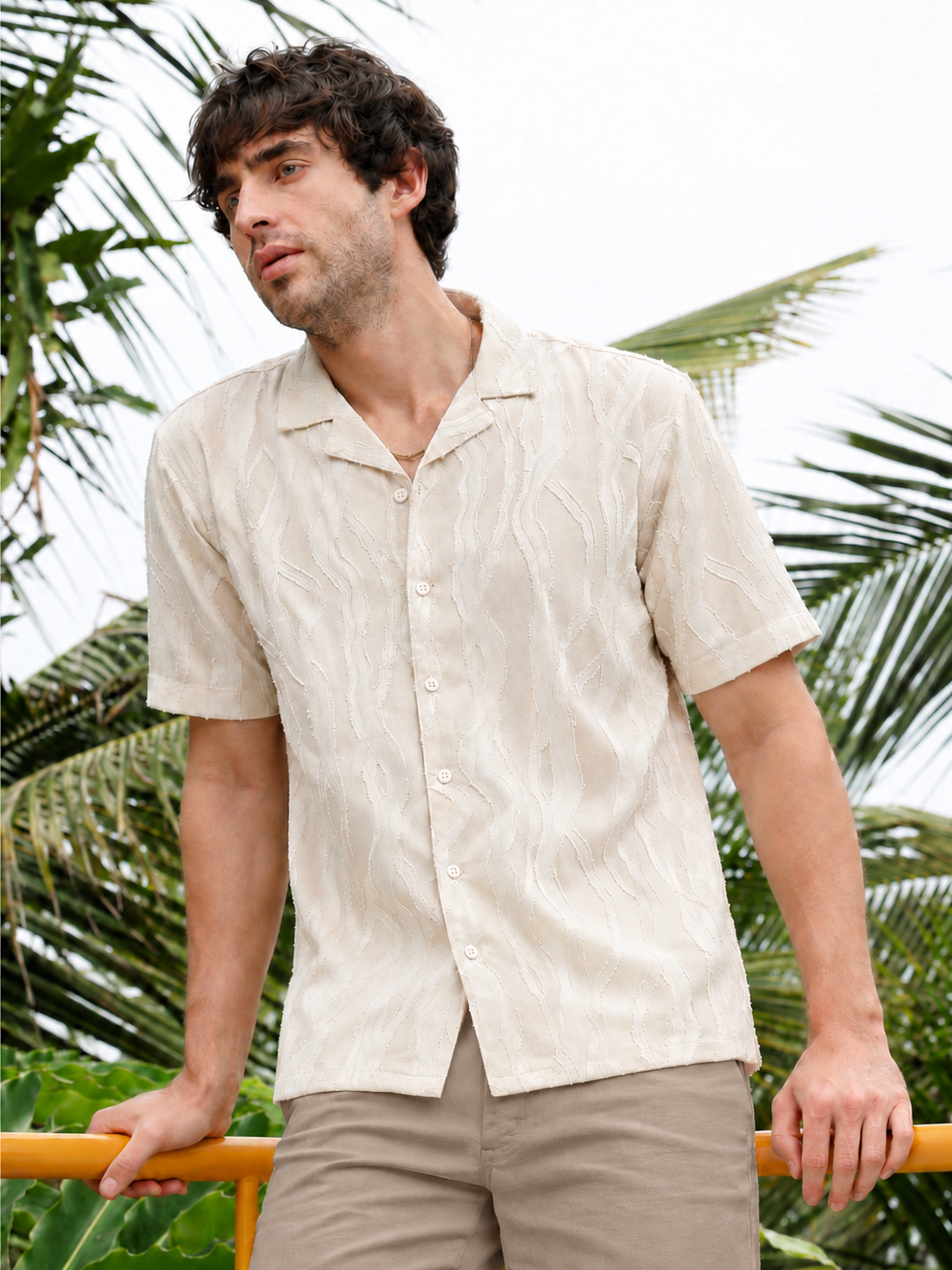 A man wearing a short-sleeve button-up camp shirt in a light beige textured fabric, standing outdoors with palm trees in the background.