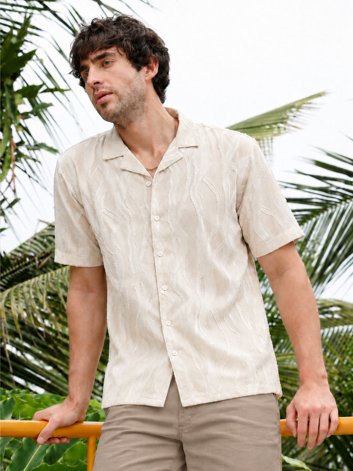 A man wearing a short-sleeve button-up camp shirt in a light beige textured fabric, standing outdoors with palm trees in the background.