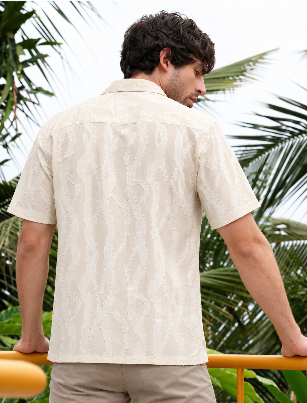 A man wearing a short-sleeve cream-colored camp shirt with a textured leaf pattern, shown from the back in an outdoor tropical setting with palm leaves.
