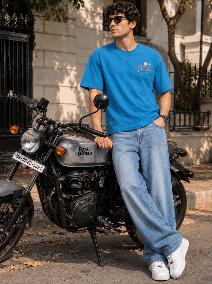 A man wearing a bright blue t-shirt with a 'Royal Enfield' logo, light blue jeans, and white sneakers leans against a silver Royal Enfield motorcycle on a street.