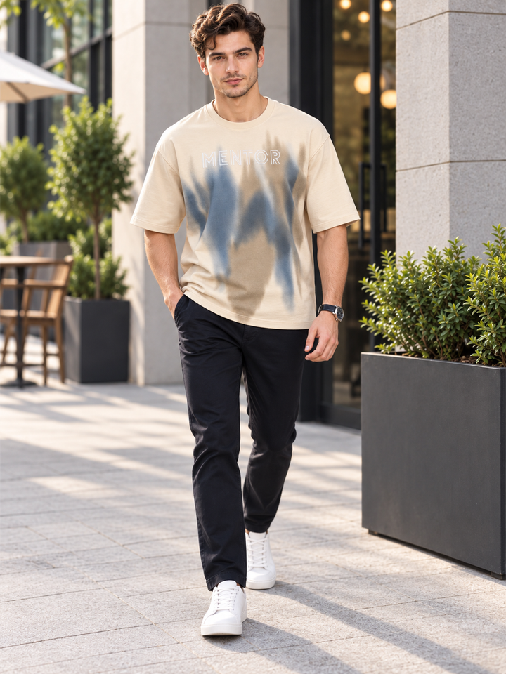 A man walking outdoors in a beige graphic t-shirt by mentor with a blue and gray abstract print, paired with black pants and white sneakers.