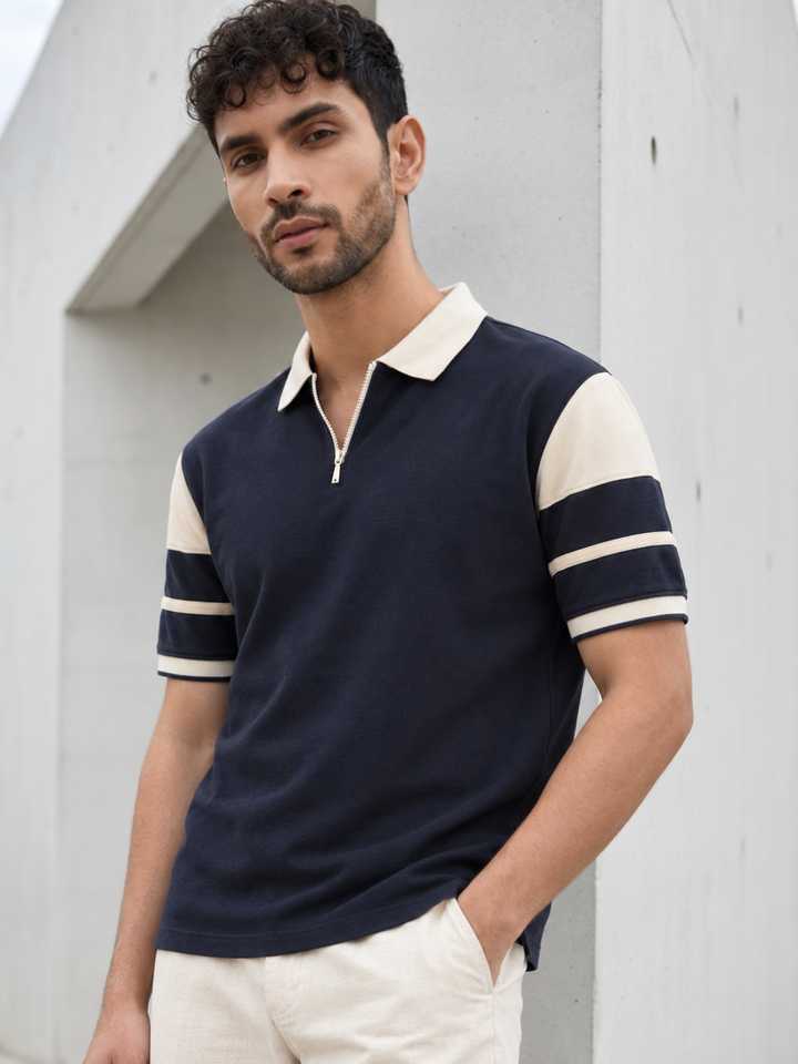 A man wearing a navy blue polo shirt with cream-colored collar and sleeve accents, paired with cream trousers, standing against a light gray concrete wall.