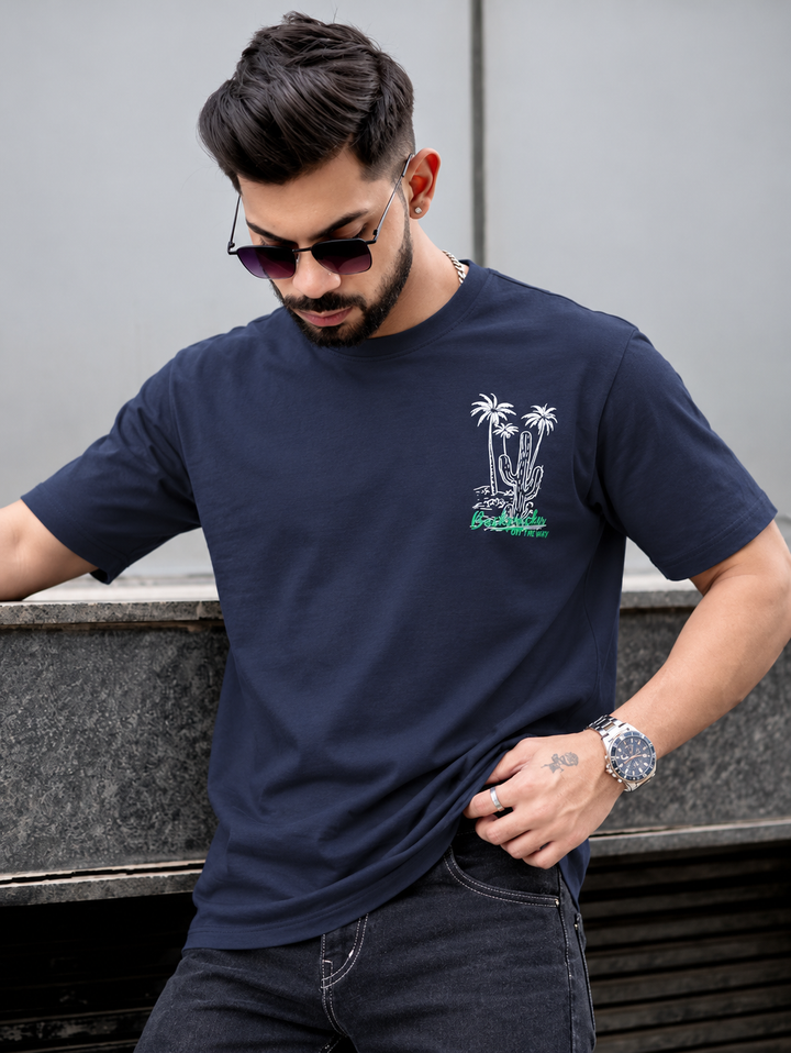 A man wearing a navy blue crewneck t-shirt with a small white palm tree graphic and green text on the left chest, paired with dark jeans and sunglasses, posing outdoors against a gray wall.