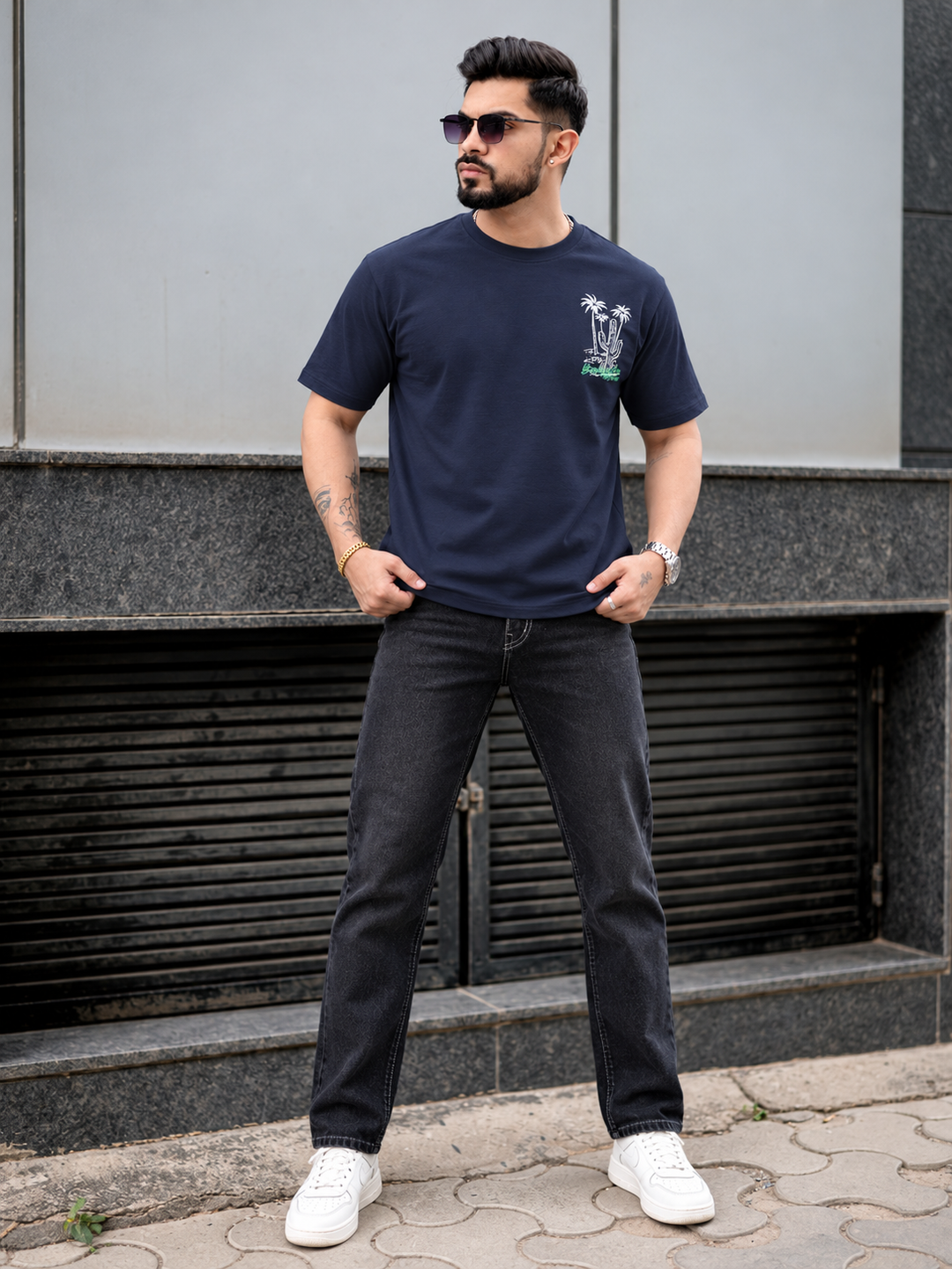A man wearing a navy blue crewneck t-shirt with a small palm tree graphic on the left chest, paired with dark gray jeans and white sneakers, standing outdoors in front of a modern gray building.