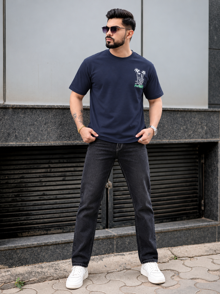 A man wearing a navy blue crewneck t-shirt with a small palm tree graphic on the left chest, paired with dark gray jeans and white sneakers, standing outdoors in front of a modern gray building.