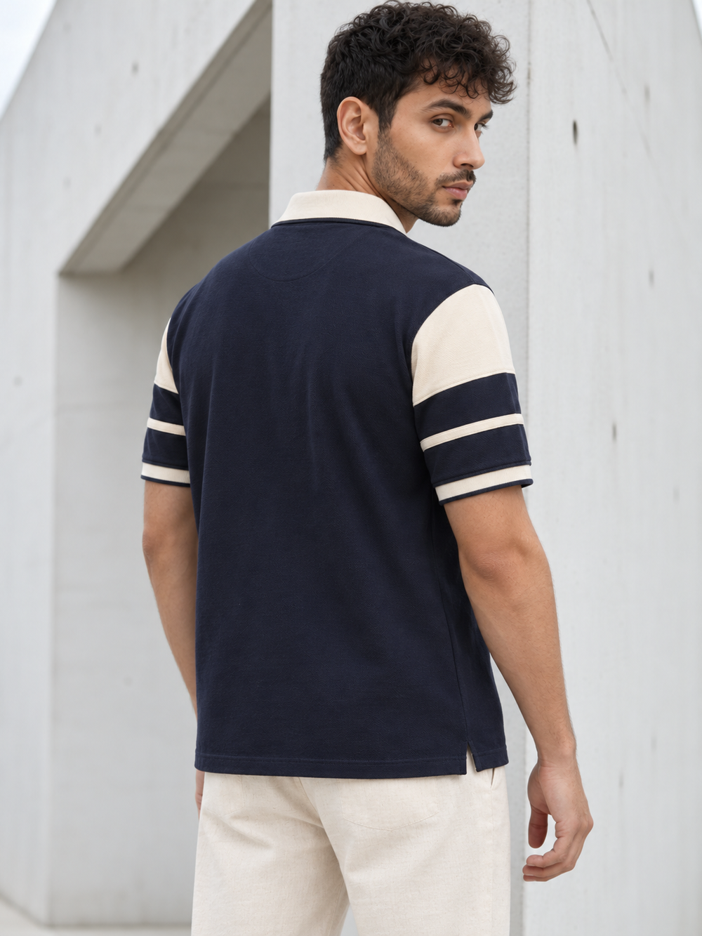 A man wearing a navy blue and cream color-blocked short-sleeve polo shirt, shown from a three-quarters back view against a minimalist concrete wall.