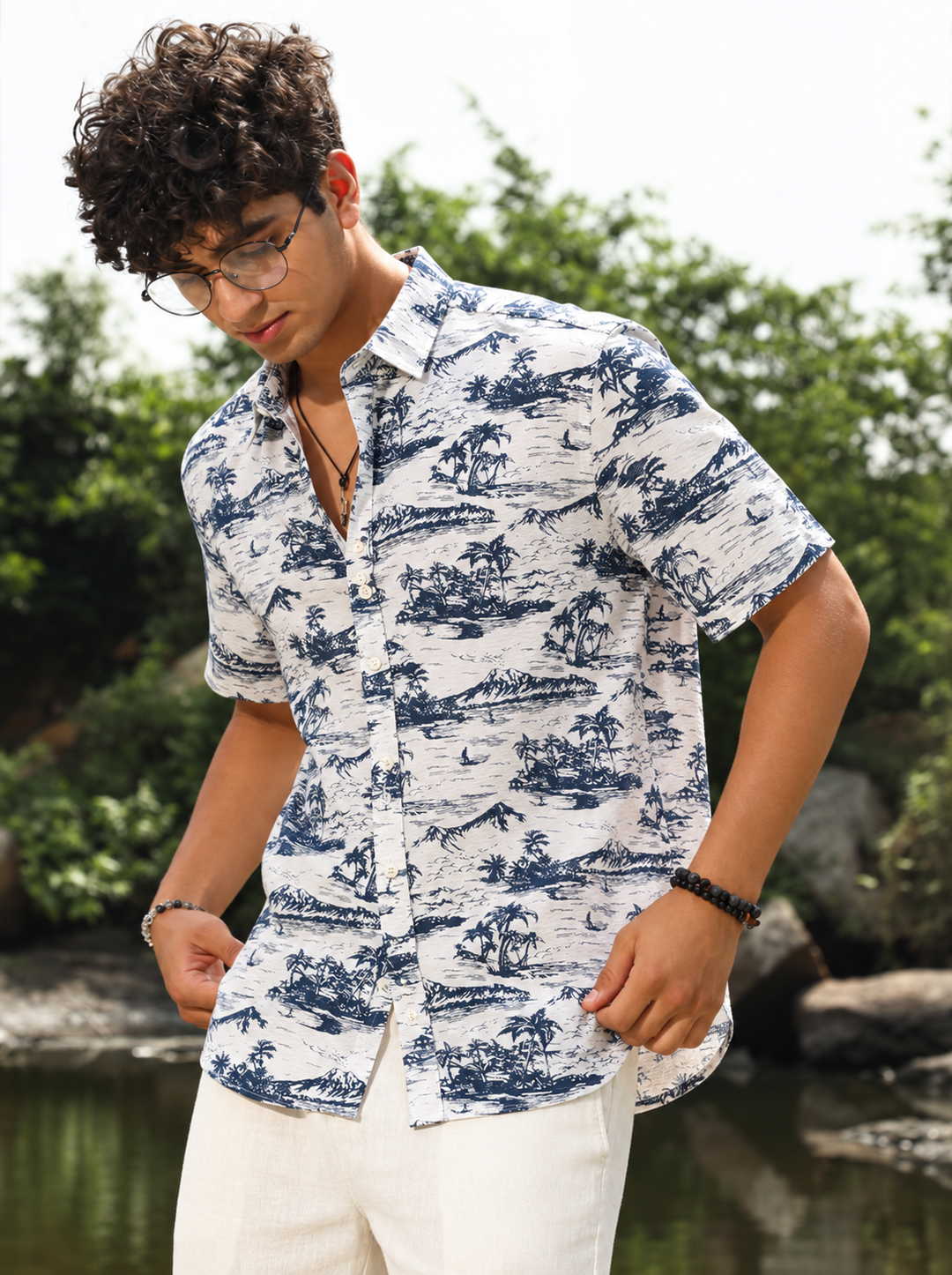 A man wearing a short-sleeve button-up shirt with a blue tropical landscape toile pattern on a white background, paired with white pants, standing outdoors near water and greenery.