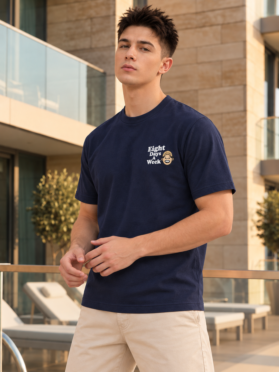 A man wearing a navy blue crewneck t-shirt with a white graphic print on the left chest that reads 'Eight Days Without' alongside a smiley face, paired with beige shorts, standing on a modern outdoor balcony.
