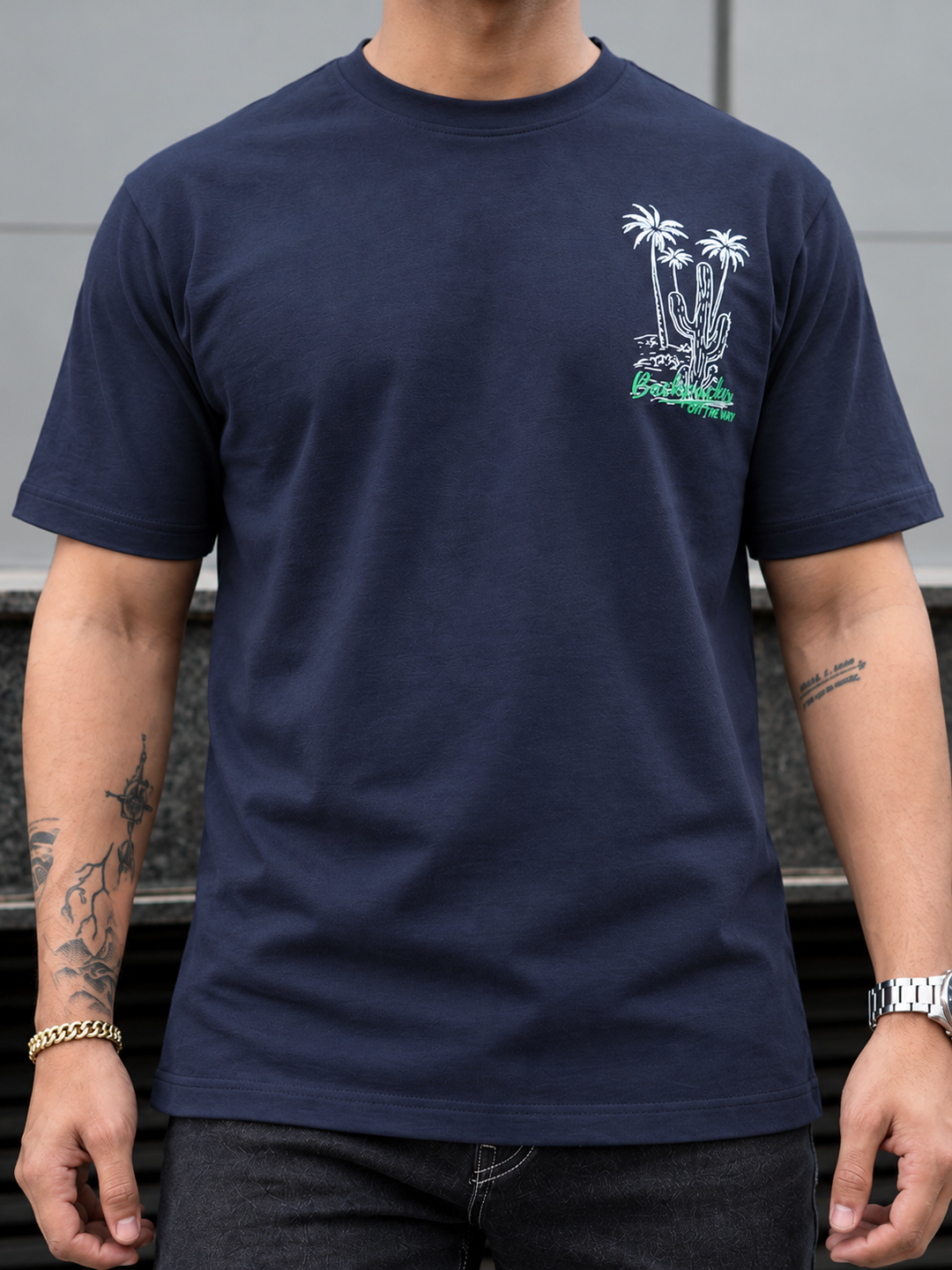 A man wearing a navy blue crewneck t-shirt with a white and green desert graphic on the left chest, featuring palm trees and a cactus.