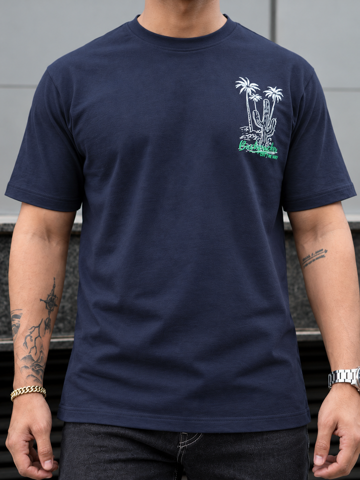 A man wearing a navy blue crewneck t-shirt with a white and green desert graphic on the left chest, featuring palm trees and a cactus.