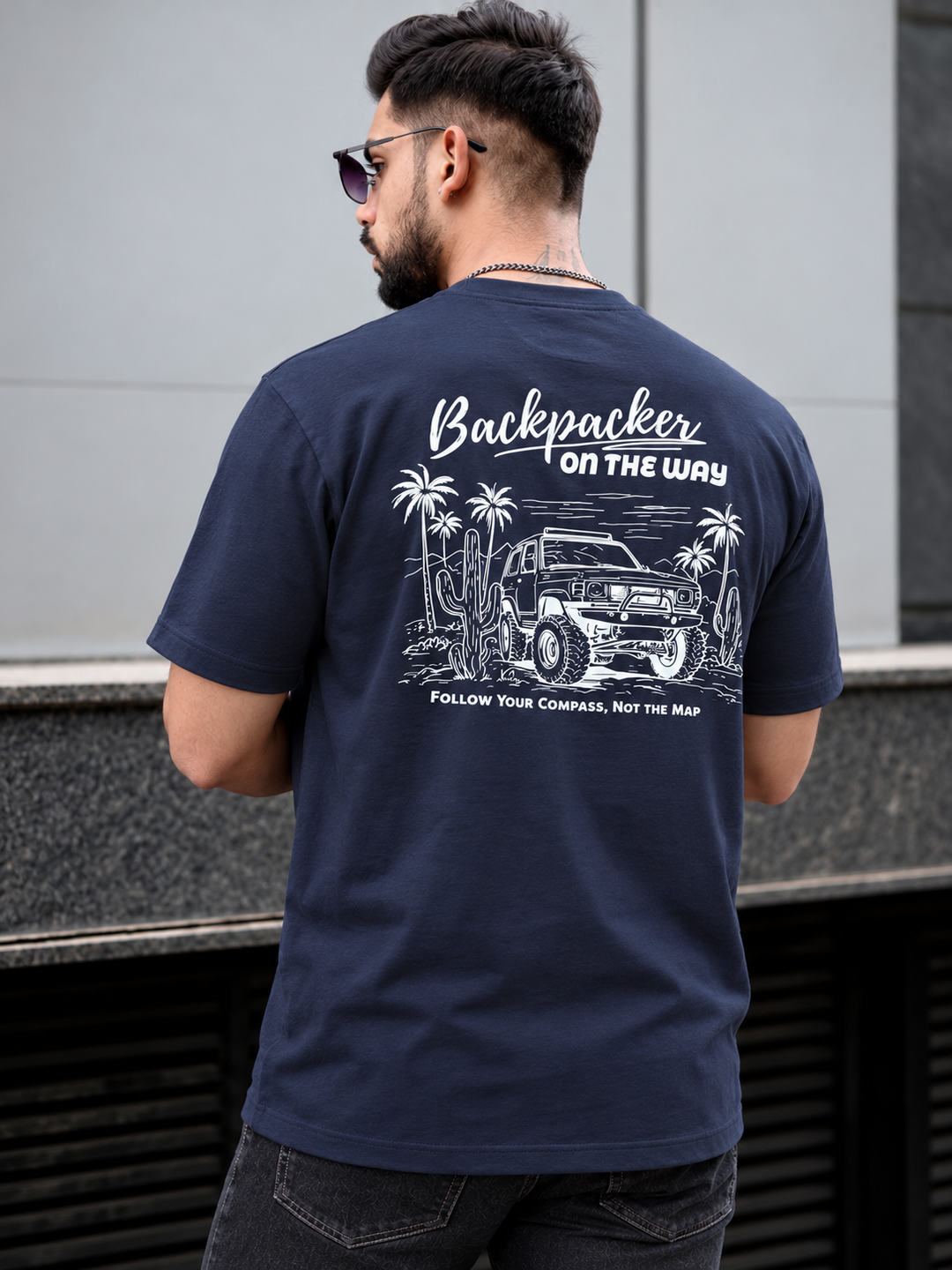 A man wearing a navy blue t-shirt with a white graphic print on the back, featuring a jeep, palm trees, and the text 'Backpacker ON THE WAY' and 'FOLLOW YOUR COMPASS, NOT THE MAP'. He is photographed from behind in an outdoor setting.