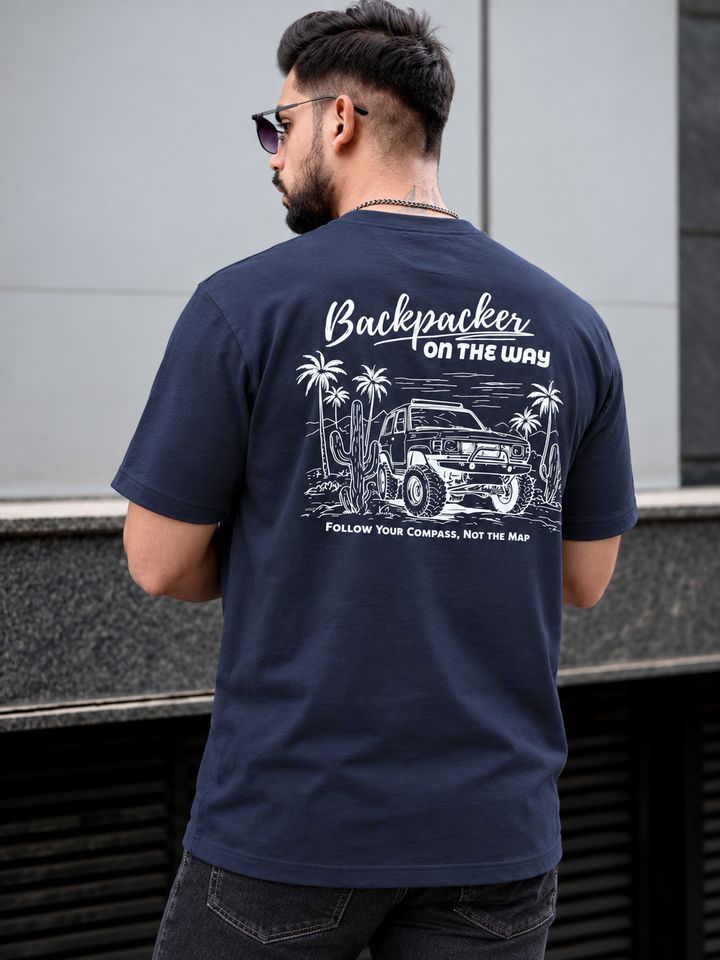 A man wearing a navy blue t-shirt with a white graphic print on the back, featuring a jeep, palm trees, and the text 'Backpacker ON THE WAY' and 'FOLLOW YOUR COMPASS, NOT THE MAP'. He is photographed from behind in an outdoor setting.