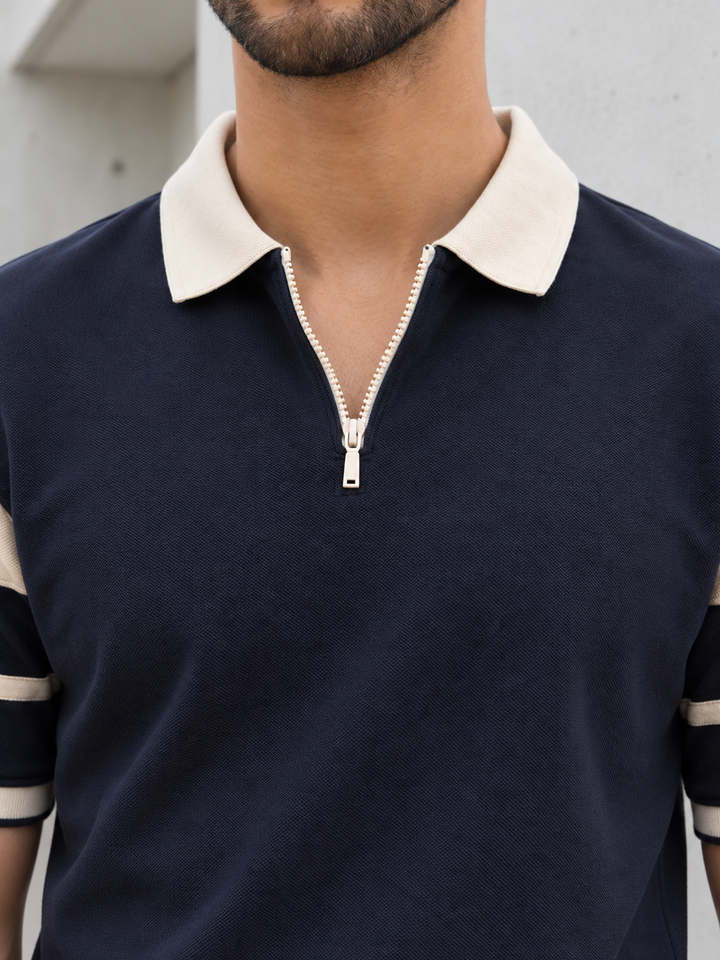 A close-up of a man wearing a navy blue polo shirt with a cream-colored collar, a half-zip neckline, and cream-and-navy striped sleeve cuffs.