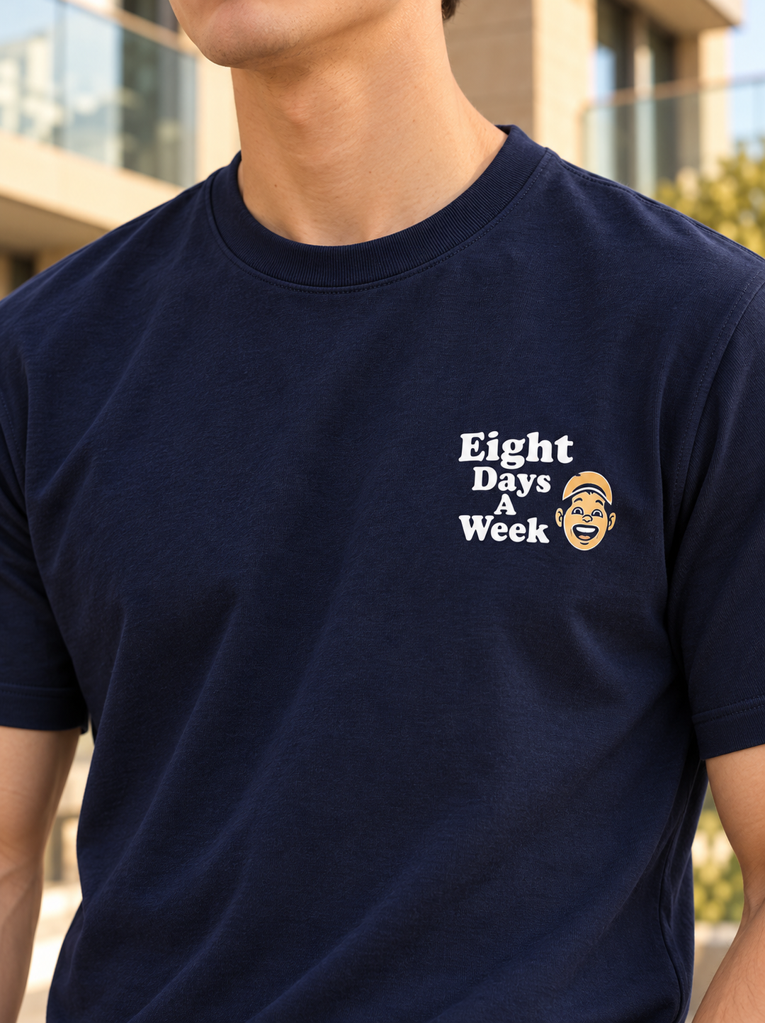 A man wearing a navy blue crewneck t-shirt with a white graphic text reading 'Eight Days A Week' and a small cartoon face illustration on the left chest, set against a blurred outdoor background.