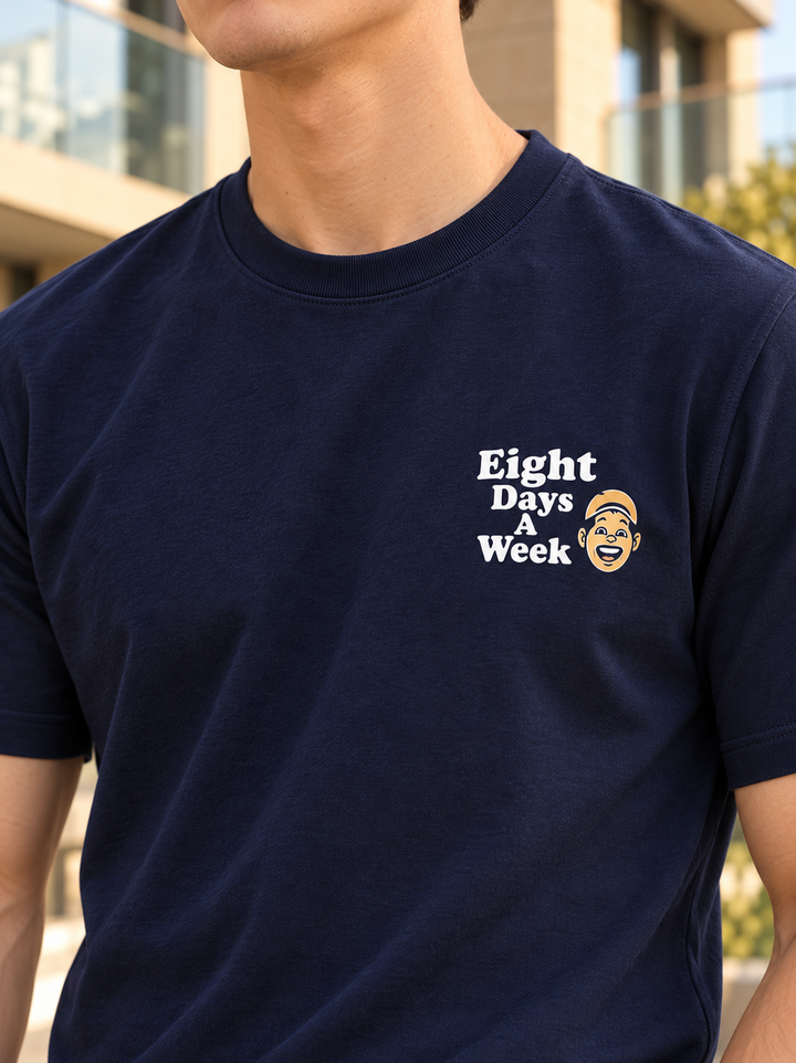 A man wearing a navy blue crewneck t-shirt with a white graphic text reading 'Eight Days A Week' and a small cartoon face illustration on the left chest, set against a blurred outdoor background.