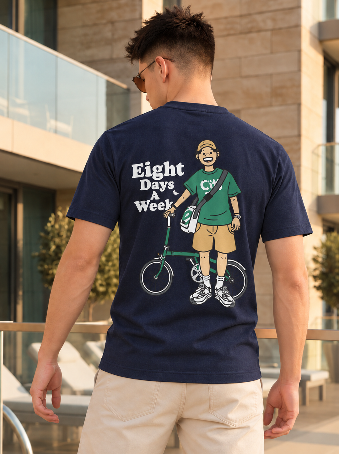 A man wearing a navy blue t-shirt with a large graphic print on the back, standing outdoors in front of a modern building. The graphic features a cartoon illustration of a man with a bicycle and the text 'Eight Days & Week'.