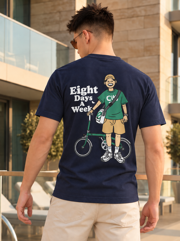 A man wearing a navy blue t-shirt with a large graphic print on the back, standing outdoors in front of a modern building. The graphic features a cartoon illustration of a man with a bicycle and the text 'Eight Days & Week'.