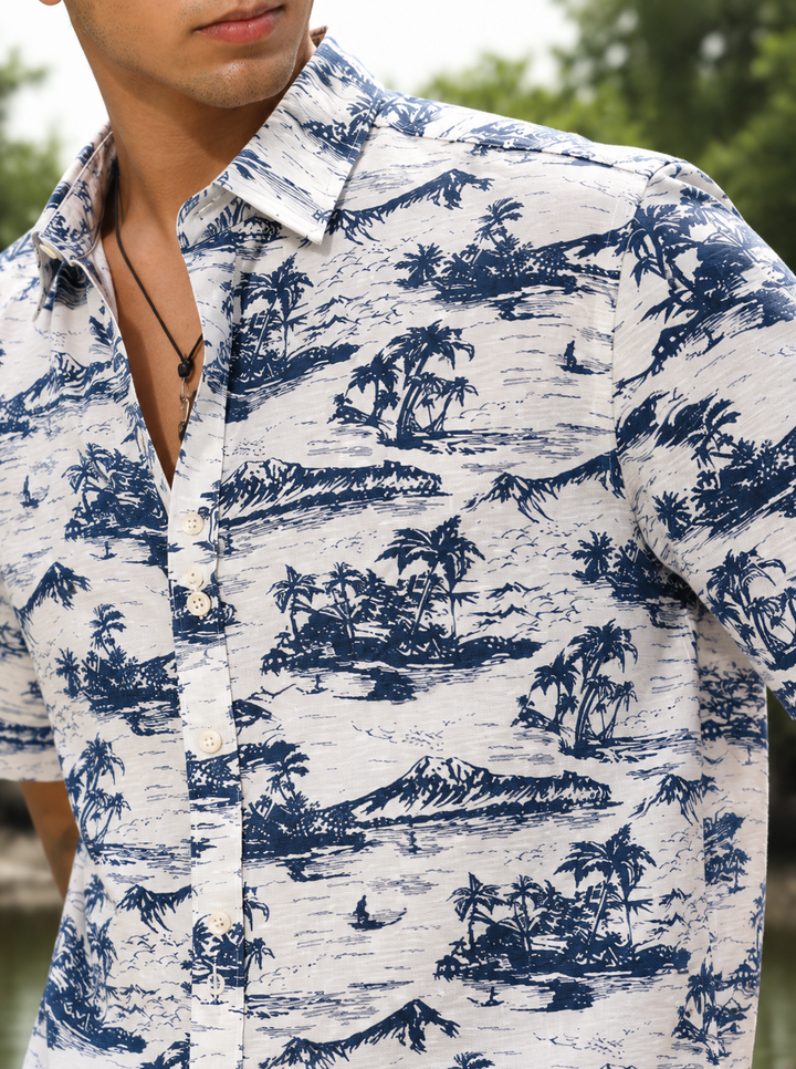 A man wearing a short-sleeve button-up shirt with a blue and white tropical island print, featuring palm trees, mountains, and waves.
