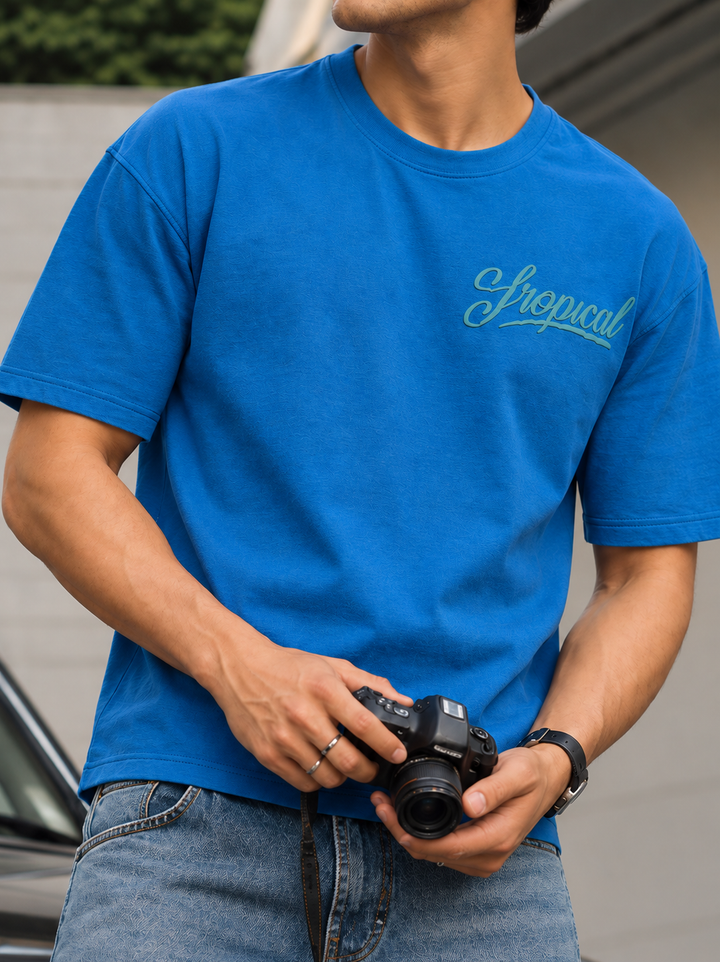 A man wearing a bright blue mentor crewneck t-shirt with a light blue cursive 'Tropical' logo on the left chest, holding a black camera. The background is a blurred outdoor setting.