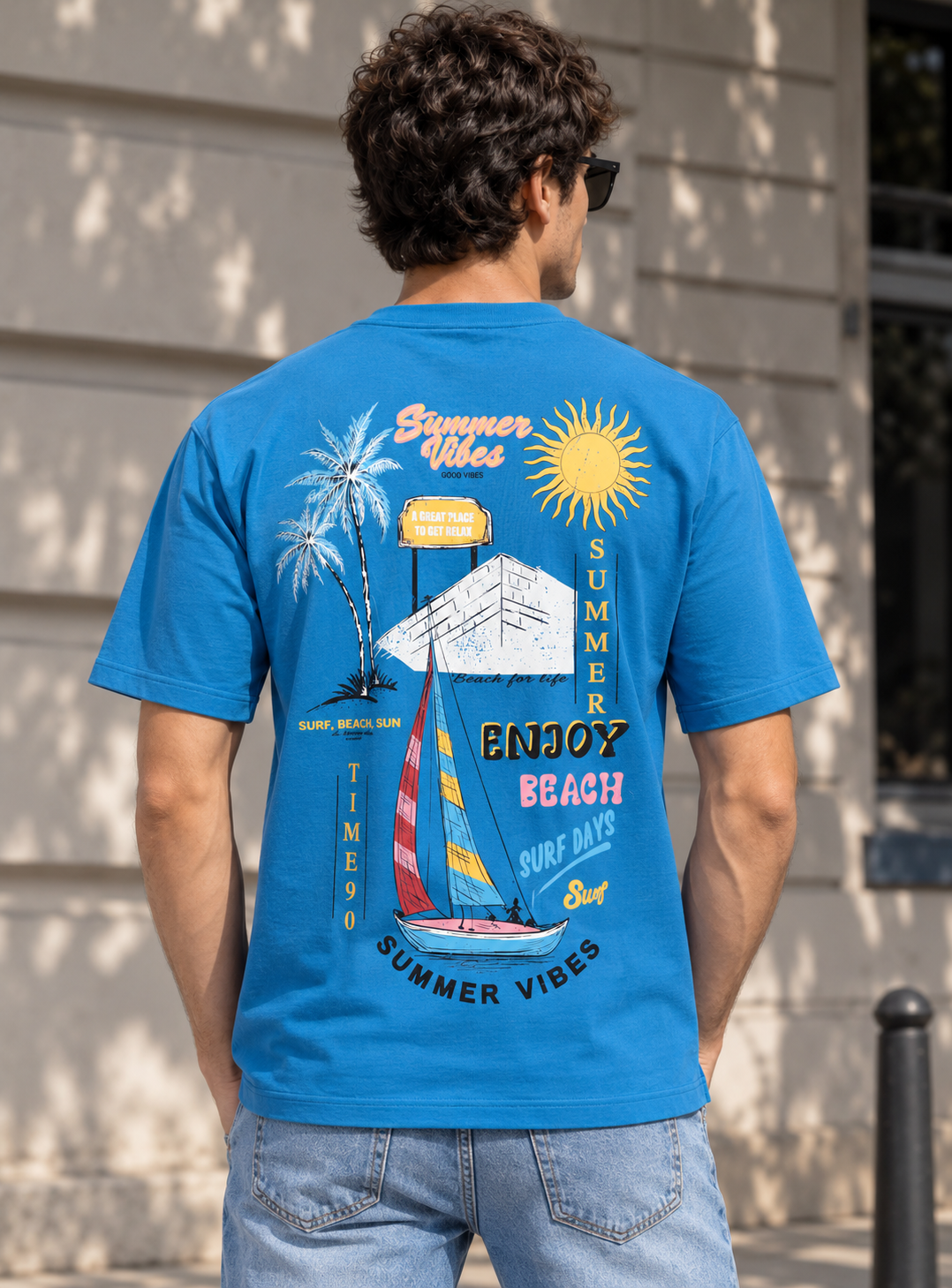 A man viewed from behind wearing a bright blue graphic t-shirt with a colorful summer beach and sailboat design, paired with light-wash jeans, standing outdoors against a beige building wall.