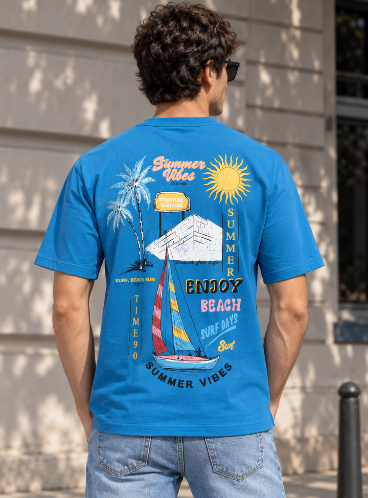 A man viewed from behind wearing a bright blue graphic t-shirt with a colorful summer beach and sailboat design, paired with light-wash jeans, standing outdoors against a beige building wall.