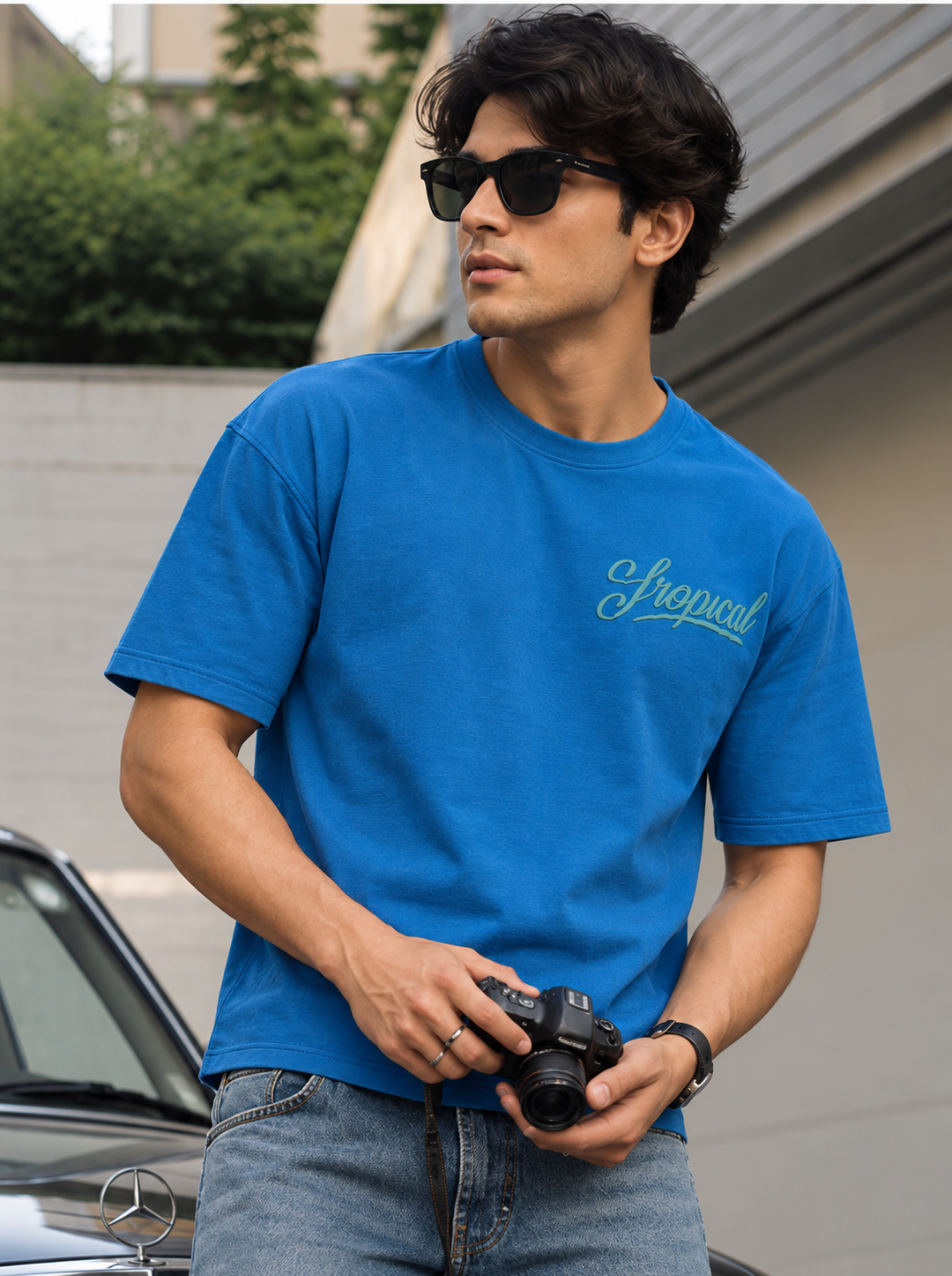 A man wearing a bright blue crewneck t-shirt by mentor with a light blue script logo on the chest, paired with blue jeans and black sunglasses, holding a camera outdoors.