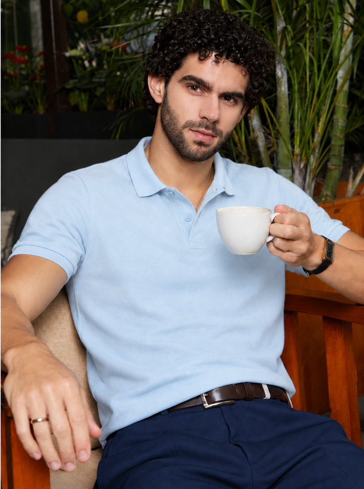 A man with curly dark hair and a beard sits in a wooden chair, wearing a light blue short-sleeve polo shirt and navy trousers while holding a white ceramic coffee mug.