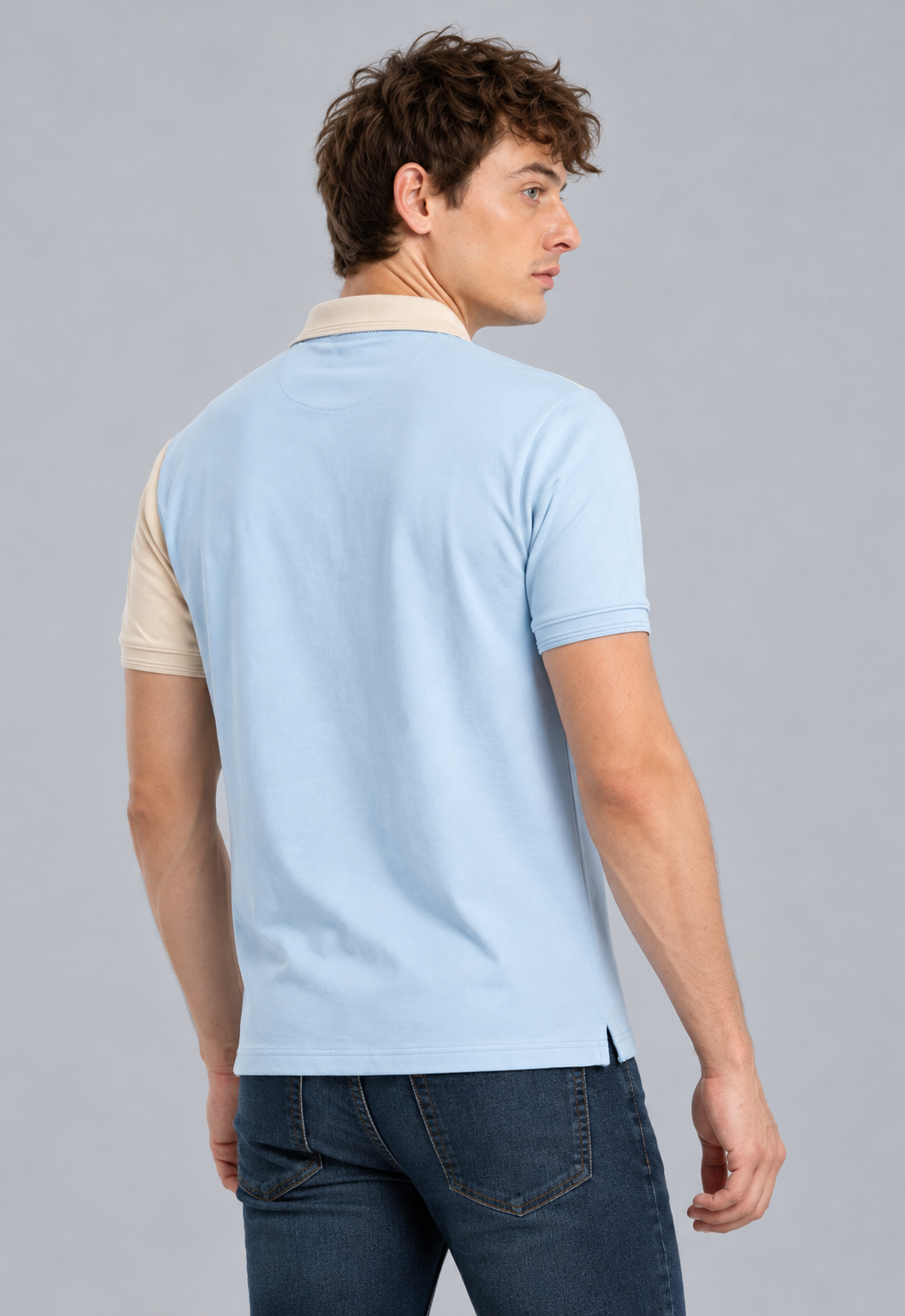 A man wearing a light blue short-sleeve polo shirt with contrasting beige sleeves and collar, paired with dark blue jeans, shown from a three-quarters back view against a solid gray background.