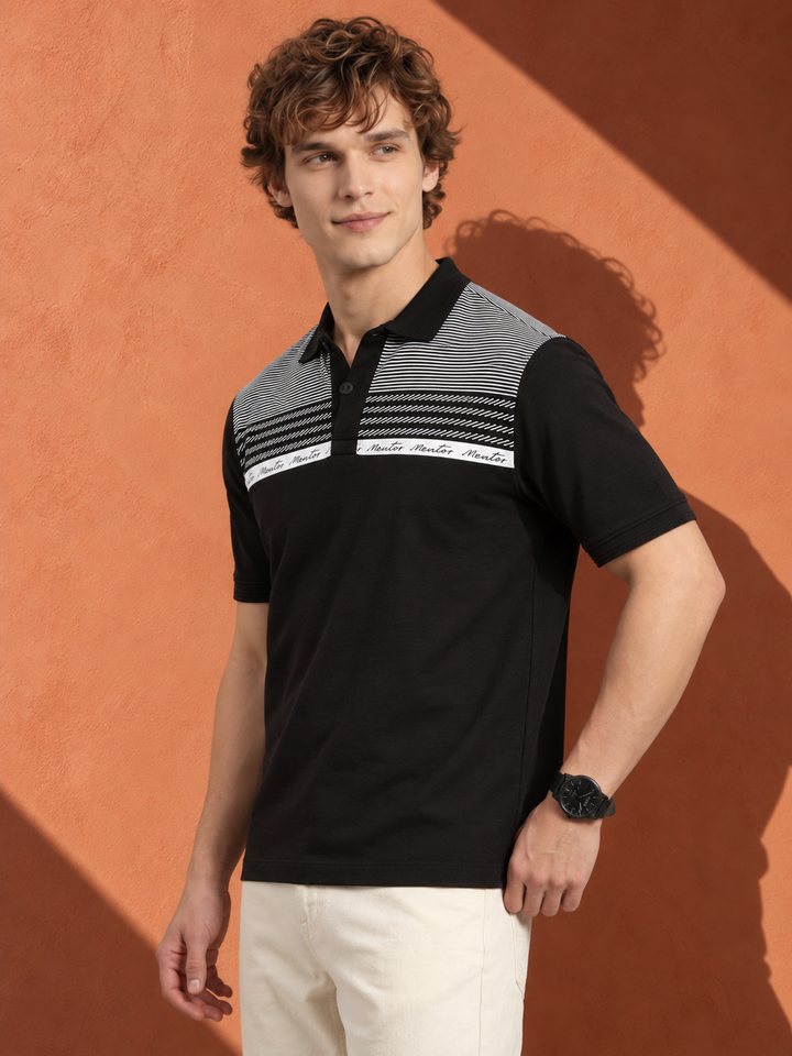A man wearing a black short-sleeve polo shirt with gray and white patterned shoulder and chest panels, paired with off-white trousers, standing against a textured terracotta wall with strong sunlight and shadows.