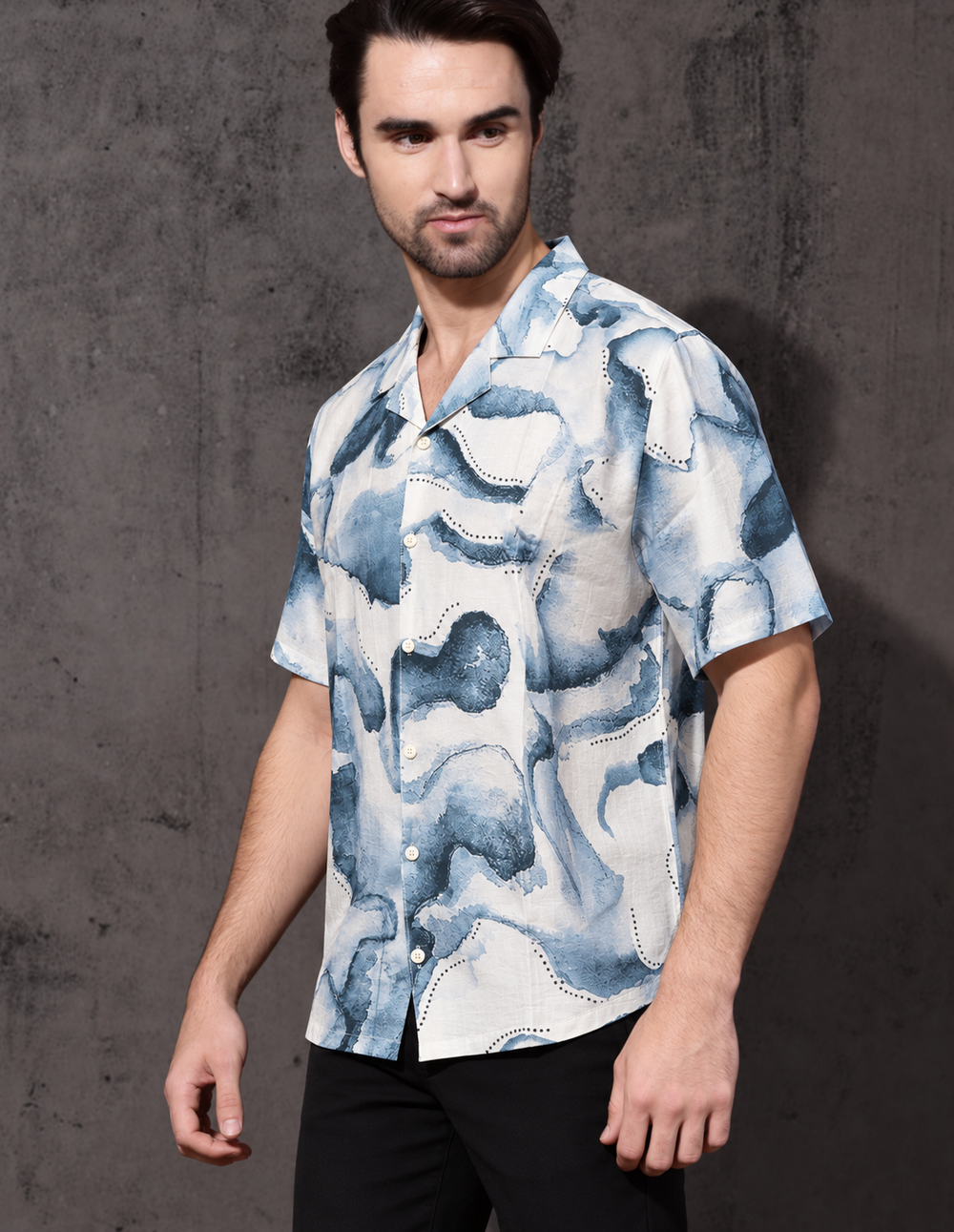 A man wearing a short-sleeve button-up shirt with a blue and white abstract watercolor pattern, paired with black pants, standing against a textured gray wall.
