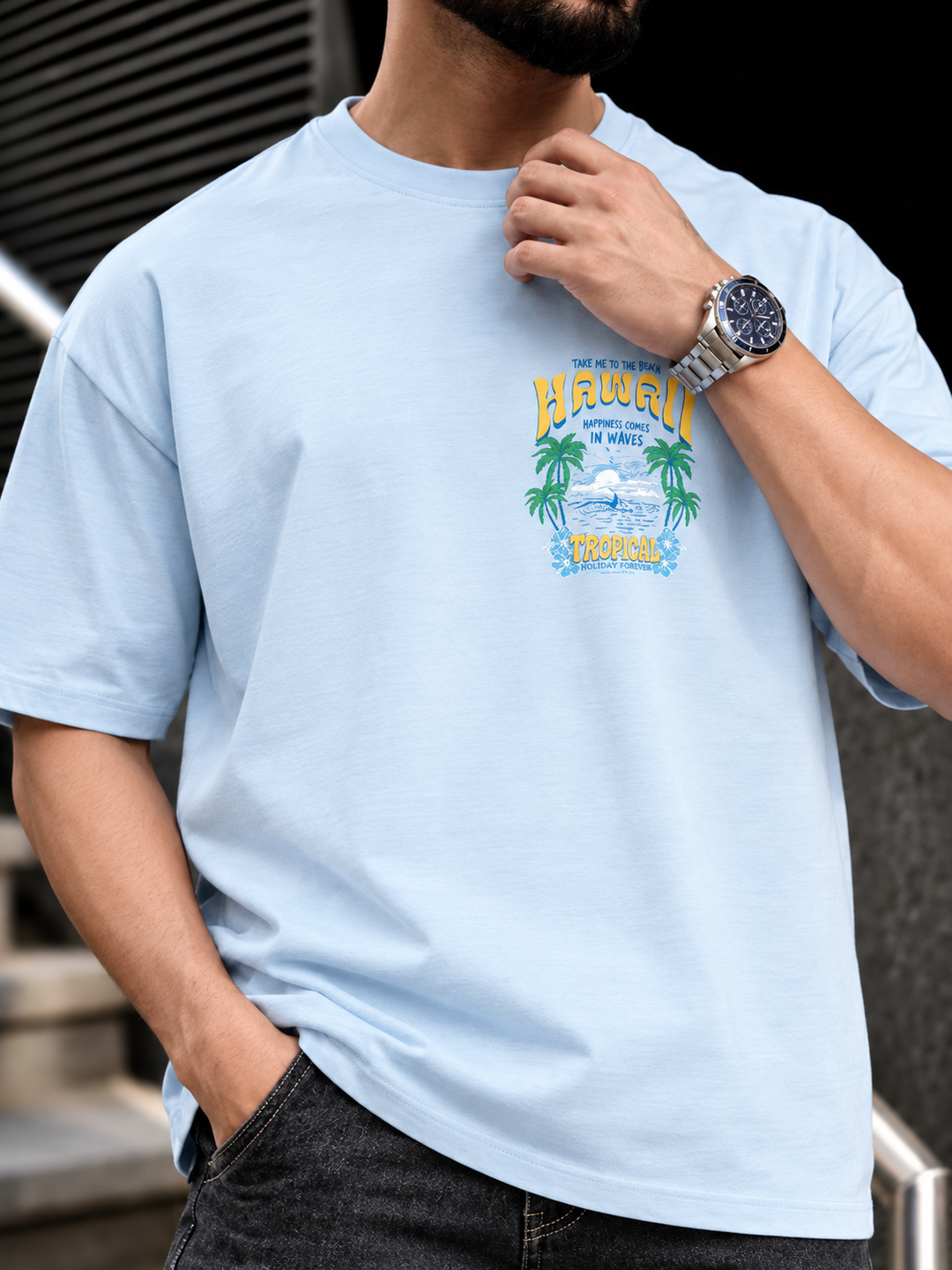 A man wearing a light blue crewneck t-shirt with a colorful graphic on the chest that reads 'HAWAII' and 'Koolohe Bay'.