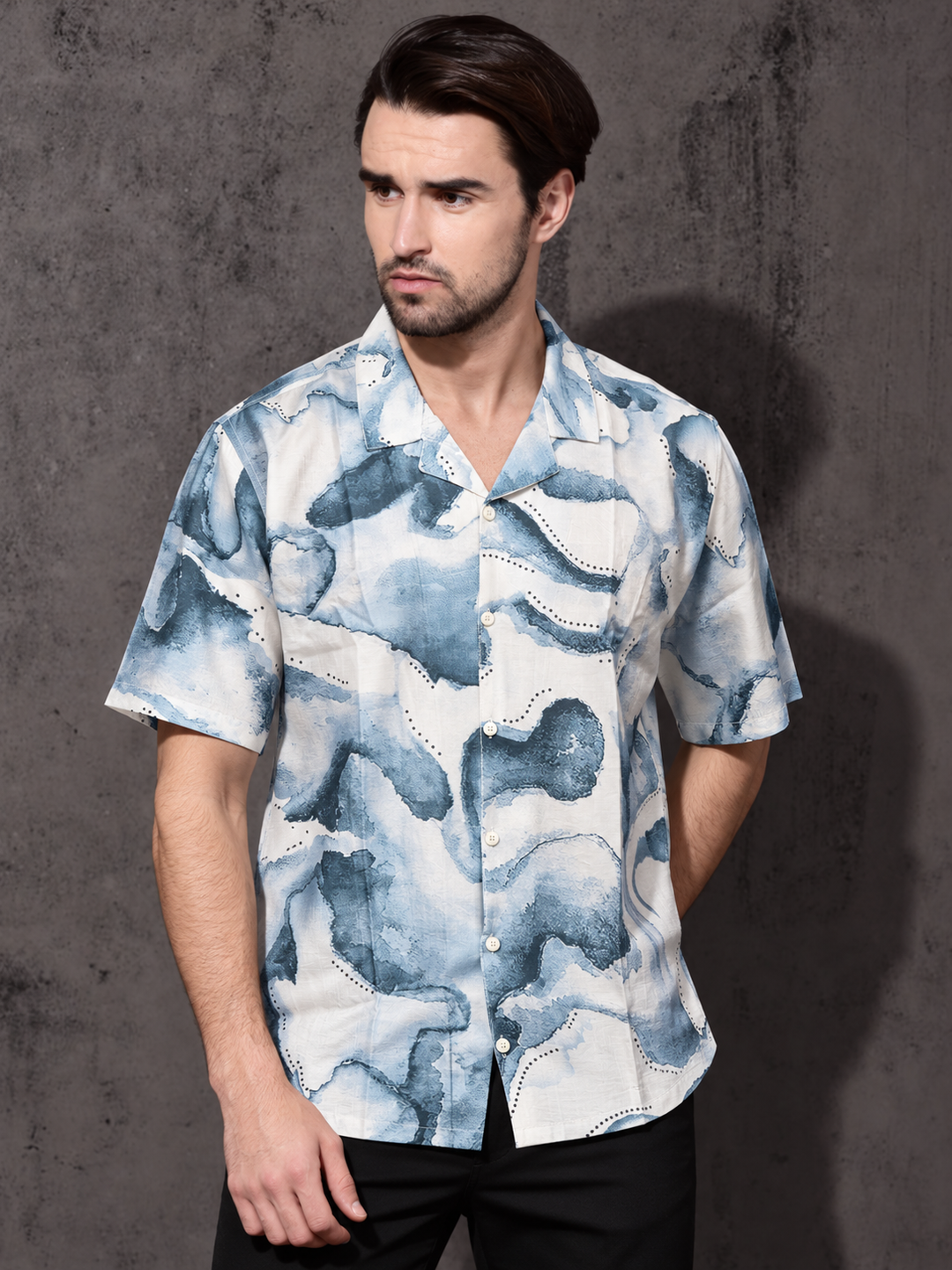 A man wearing a short-sleeve button-up shirt with a blue and white abstract watercolor pattern, posed against a textured gray wall.