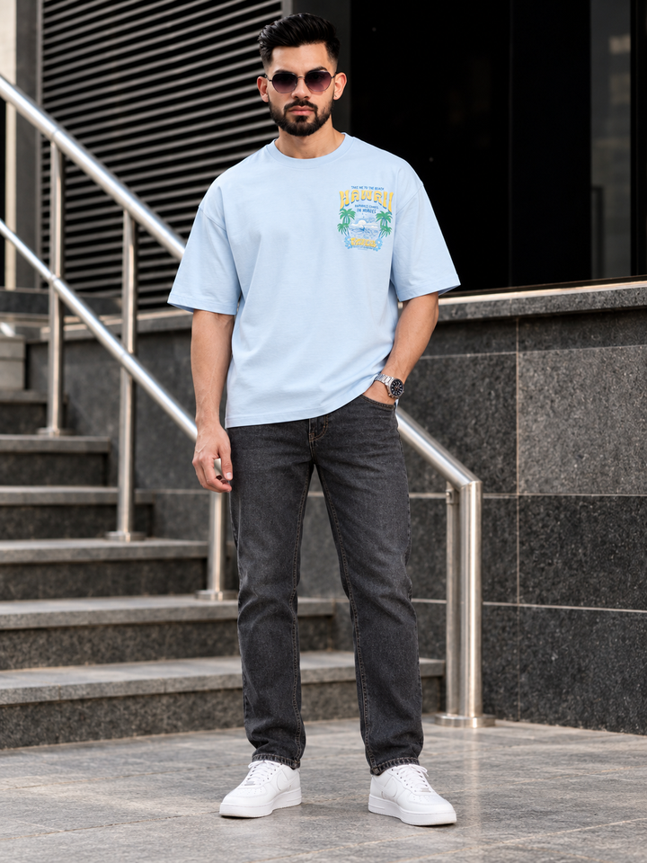 A man wearing a light blue graphic t-shirt, dark gray jeans, and white sneakers stands in front of outdoor stone stairs with metal railings.