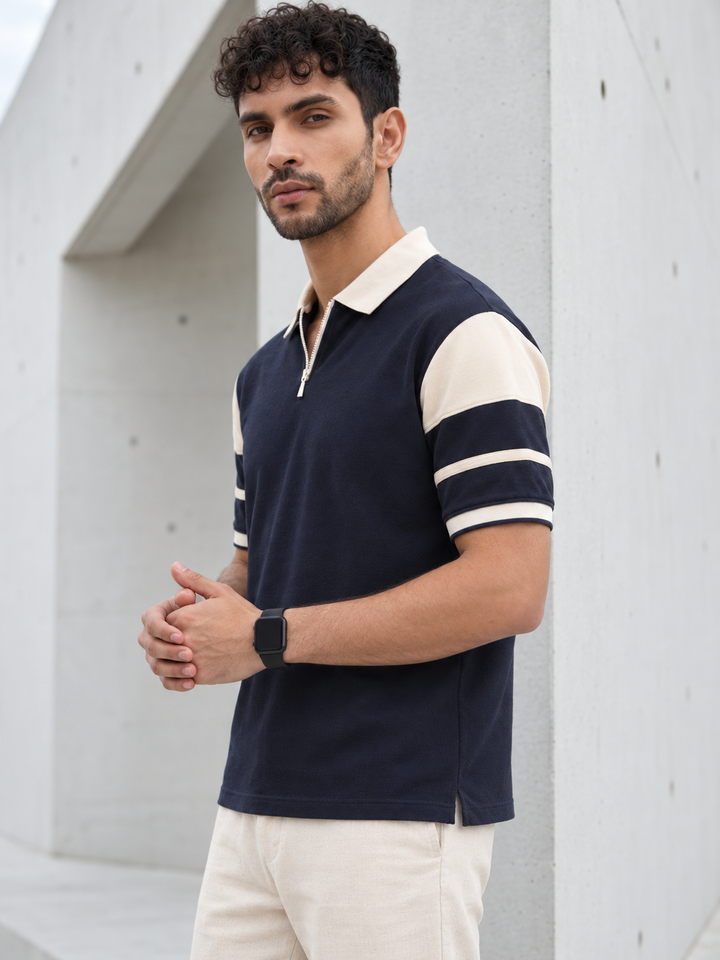 A man wearing a navy blue polo shirt with cream collar and sleeve stripes, paired with cream pants, standing against a modern concrete wall.