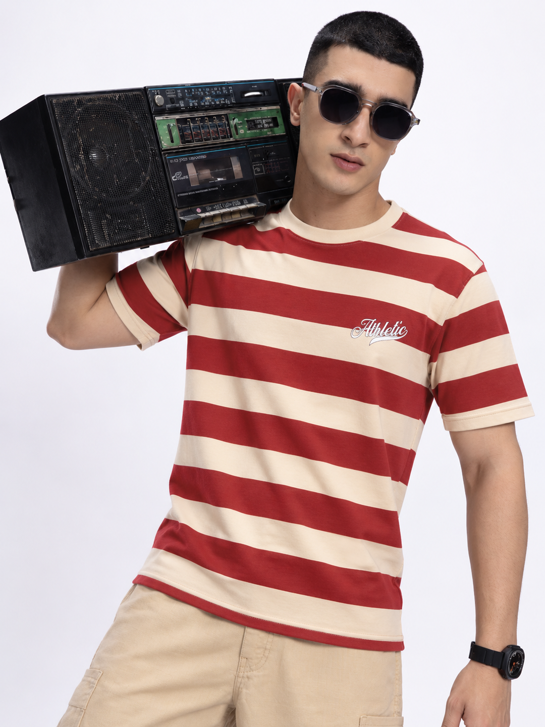 A man wearing a red and beige horizontally striped t-shirt with a cursive 'Athlete' logo on the chest, holding a vintage boombox over his shoulder, set against a plain white studio background.