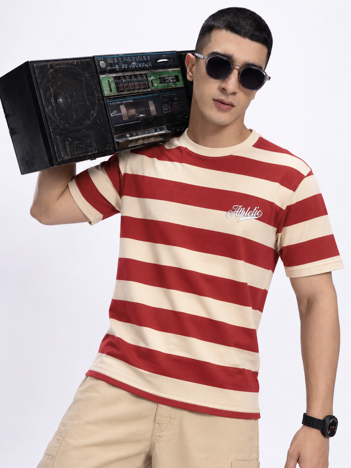 A man wearing a red and beige horizontally striped t-shirt with a cursive 'Athlete' logo on the chest, holding a vintage boombox over his shoulder, set against a plain white studio background.