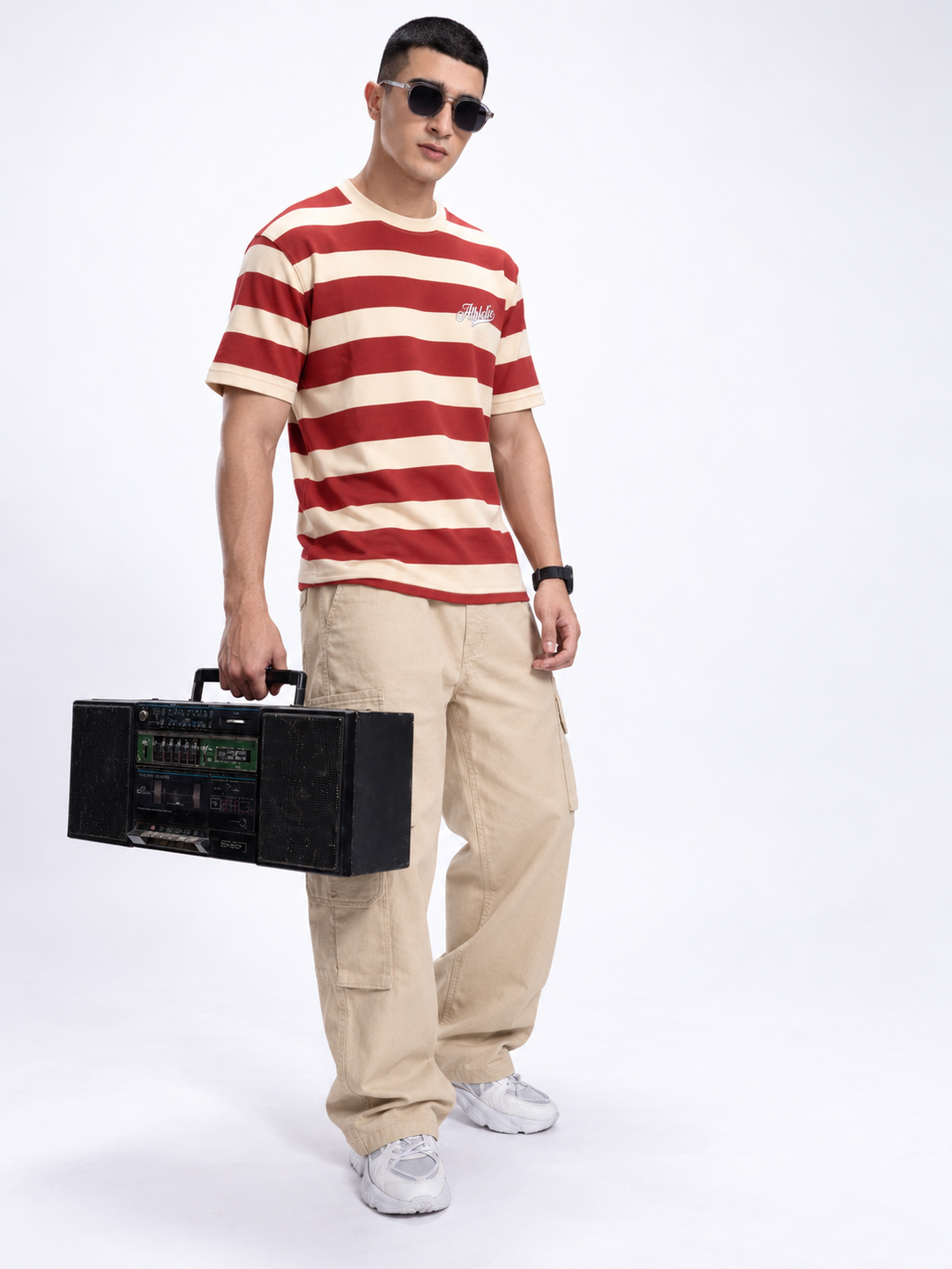 A man wearing a red and beige striped t-shirt, beige cargo pants, and white sneakers holds a vintage black boombox against a plain white studio background.