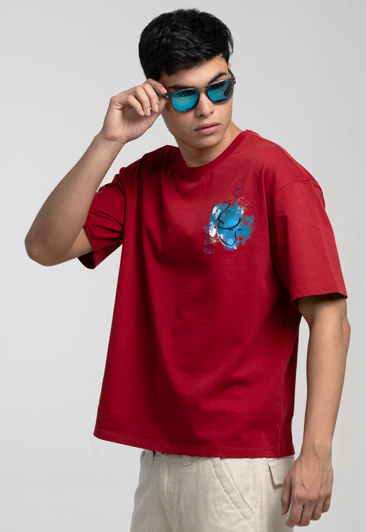 Person wearing a red t-shirt with a blue graphic design, holding sunglasses, on a plain background