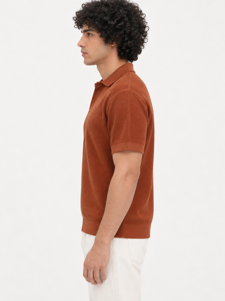 A man in side profile wearing a rust-orange short-sleeve knit polo shirt and white pants, photographed against a clean white studio background.